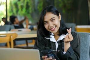 cheerful asian woman holding smartphone and show korean love signs while working with laptop remotely from cafe photo