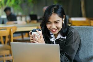 emocionado asiático mujer bebida café y utilizando ordenador portátil a tener en línea reunión en cafetería, digital nómada foto