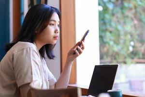 pensive asian woman sitting near window with laptop on wood table holding smartphone looking away reflecting on wearing stylish casual clothes photo