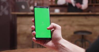 uomo scorrimento su un' Telefono con verde schermo su il backgorund di un' Vintage ▾ pub, ristorante o caffè negozio video