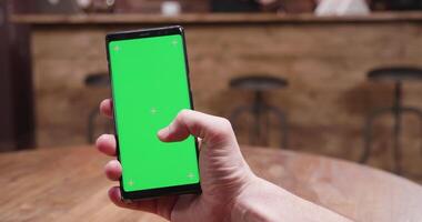 Swiping on a phone with green screen in a vintage cafe. Man holds a modern smartphone video