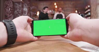 pOV antal fot av manlig händer spelar en video spel på en tabell i en årgång Kafé medan de bartender är talande med en kund i de bakgrund på de bar disken.