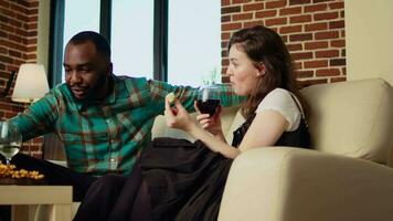 Interracial couple at social gathering talking on couch while enjoying glass of wine. Husband and wife guests at apartment party having pleasant conversation in brick wall home, eating snacks video