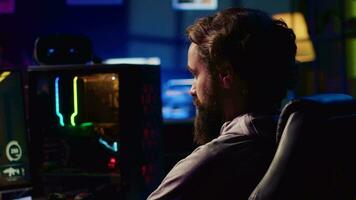 Man in dark living room playing video games on gaming PC at computer desk during leisure time. Gamer battling enemies in online multiplayer shooter from neon lights ornate home