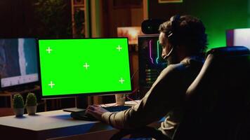 Man in dimly lit apartment playing video games on green screen gaming PC at computer desk, enjoying day off from work. Gamer battling enemies in online multiplayer shooter on mockup monitor