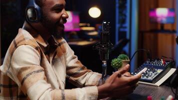 African american man interviewing celebrity, reviewing notes to ask pertinent questions. Journalist doing radio interview in apartment studio, wearing headphones and talking on mic with guest video