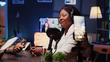 Journalist records interview with cellphone on tripod stabilizer in studio, reviewing notes to ask quality questions to guest. Close up of woman using phone to film vlog, talking on mic with celebrity video