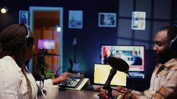 Vlogger show host adjusting analog mixer while talking with guest in neon lights ornate personal studio, ensuring perfect sound quality. Man and woman filming online podcast episode, zoom out shot video