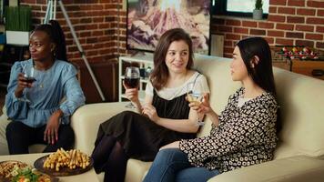 multi-etnisch Dames zittend Aan bankstel in knus appartement leven kamer drinken wijn, chatten. verschillend groep van meisjes verzameld Bij huis voor speciaal gelegenheid, bespreken over hun vriendjes video