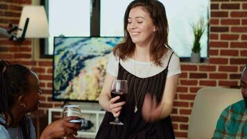 Woman chatting with diverse apartment party guests during weekend gathering, being amused while gossiping about friend. Multiracial group of people listening to host sharing funny story video