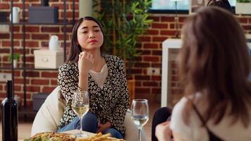 Asian woman at apartment party paying attention to interesting story shared by friend while eating gourmet cheese from charcuterie platter. BIPOC guest listening to group of people chatting video