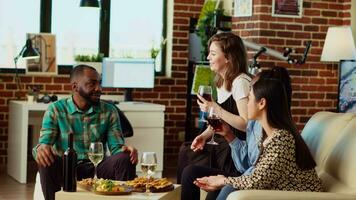 multiracial groupe de gens lancement anniversaire fête pour leur ami, en buvant de l'alcool et en mangeant de charcuterie planche. joyeux invités en riant dans moderne vivant pièce après cadeau hôte présente video