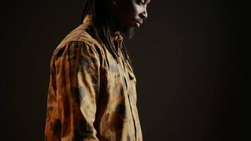 Trendy cool guy smiling in front of studio camera and posing, feeling confident with hair dreads and camo clothing items. Person being cheerful, modern fashion style over backdrop. Handheld shot. video