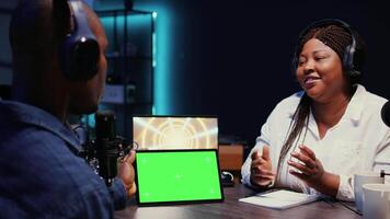 Woman invited to podcast watching internet clips on green screen tablet with host in studio, discussing content. African american guest reacting to videos on mockup device during online comedy show