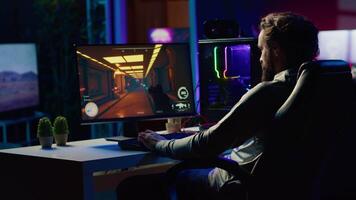 Man in dark living room playing videogame on gaming PC at computer desk, relaxing by shooting enemies. Gamer battling flying robots in online singleplayer shooter from neon lights ornate home video