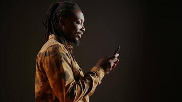 moderne la personne navigation en ligne les pages Web sur mobile téléphone, vérification le sien réseaux sociaux pour des textes et posant dans studio. africain américain homme en utilisant téléphone intelligent applications à rester connecté avec amis. video