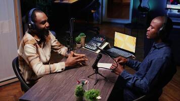 Vlogger show host interviewing guest in neon lights ornate personal studio, discussion about his life. BIPOC men filming online podcast episode, talking with each other, jib down shot video