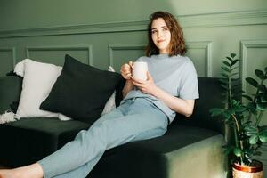 Relaxed Young Woman Reflecting with Tea Coffee in Home Setting. Serene Mood and Introspection in Cozy Indoor Ambiance. Confident Solitude and Peaceful Demeanor in Tranquil Surroundings. photo