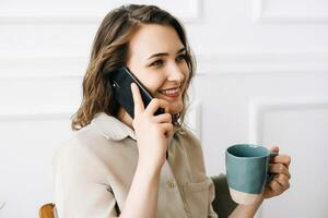 Happy Young Woman Enjoying a Phone Call While Savoring a Drink in Leisure Time. Joyful Young Woman Talking on Phone, Enjoying a Relaxing Moment with a Cup of Drink in Her Free Time photo