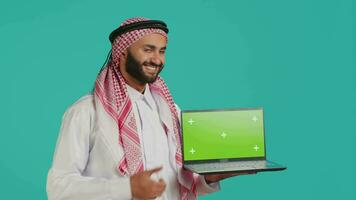 Islamitisch persoon shows groene scherm laptop, presenteren modern geïsoleerd lay-out voor kopieerruimte. jong volwassen presentatie van blanco Chroma sleutel mockup Aan draadloze pc, vervelend thobe en hoofddoek. video