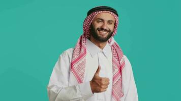 meio Oriental pessoa dá polegares acima dentro frente do Câmera, expressando aprovação e positividade enquanto ele desgasta tradicional muçulmano roupas. jovem adulto dentro nacional árabe traje, gostar placa. video