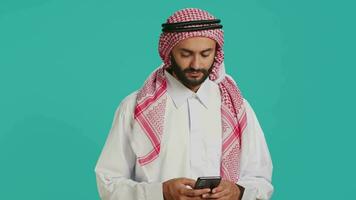 Muslim guy holding smartphone and feeling pleased after hearing good news, typing online messages on social media network. Middle eastern person scrolling internet pages, traditional clothes. video