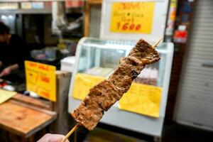 de cerca mano de turista participación y comer parilla Kobe carne de vaca madera palo en borroso parilla Kobe carne de vaca tienda a kuromon mercado, osaka, Japón. foto