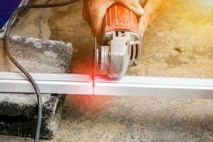 Closeup hands of laborer holding electric angle grinder working cutting aluminium lumber at construction site with sun flare background. photo