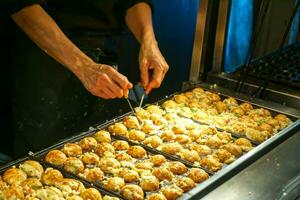 manos de cocinero Cocinando japonés bocadillo comida takoyaki preparar para rebaja a cliente en comida mercado. takoyaki es un en forma de bola hecho de un trigo a base de harina masa y cocido en un especial moldeado cacerola. foto