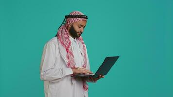 model- gebruik makend van pc touchpad in studio, gekleed in traditioneel Islamitisch kostuum en kufiyah. midden- oostelijk persoon Holding modern strak laptop en scrollen websites of communiceren online. video