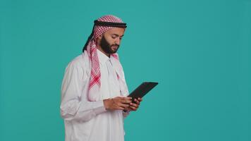 Young adult navigating on gadget in studio, browsing websites online on modern device. Muslim guy in traditional costume using tablet to check internet pages, chatting on social media. video