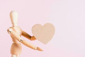 Wooden man holds out a cardboard heart in his hand on a pink background photo