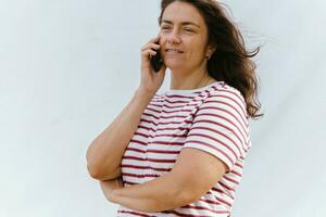 de edad mediana caucásico mujer en a rayas blusa alegremente conversando en móvil teléfono foto