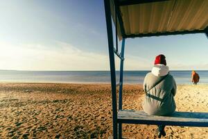 woman in an autumn jacket and hat is sitting alone on a bench on a deserted autumn beach on a sunny day photo