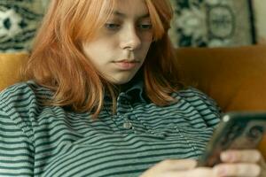 Red-Haired Teenage Girl Engrossed in Smartphone for Social Media Communication on Couch photo