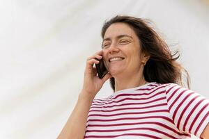 sonriente de edad mediana caucásico mujer con largo pelo hablando en Teléfono móvil en contra brillante fondo foto