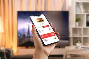 hombre pedidos un hamburguesa vía teléfono inteligente en el vivo habitación, creando un Perfecto mezcla de tecnología conveniencia y disfrute en rápido comida en frente de el televisión foto