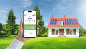 App on a mobile phone monitors the collection and consumption of electricity by solar panels. In the background is a house with solar panels on the roof photo