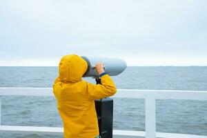 un niño en un chaqueta soportes en un blanco muelle. mirando mediante prismáticos a el mar. frío clima. espalda ver foto