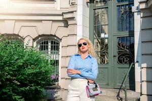 A young lady dressed in a blue shirt and white trousers is standing in the city. Blonde hair, sunglasses. a woman stands outside the office outdoors in summer, warm weather photo