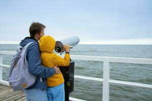 son and father look through coin binoculars at the cold sea. dad and son are standing on the pier photo