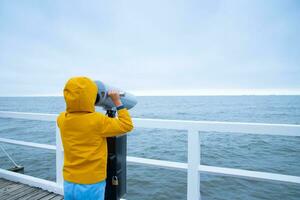 el chico mira mediante prismáticos a el báltico mar. frío otoño clima. un niño en un chaqueta soportes en el muelle y mira a el mar. foto