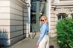un joven mujer vestido en un azul camisa y blanco pantalones es caminando mediante el ciudad. calentar verano clima. foto