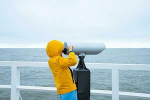 A child in a yellow jacket is standing on the pier, looking through binoculars at the sea. Cold summer weather. photo