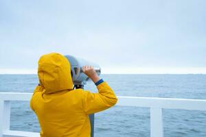 un niño en un chaqueta soportes en el muelle y mira a el mar. el chico mira mediante prismáticos a el báltico mar. frío otoño clima. foto