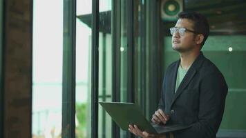 A handsome asia young stands window side by the window, he is dressed in a black suit, an office worker. video