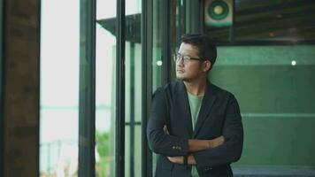 A handsome asia young stands window side by the window, he is dressed in a black suit, an office worker. video