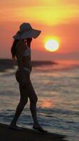 Unrecognizable woman in white bikini, sun hat walking on beach at sunrise on background solar disk video