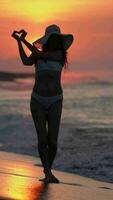 Woman in bikini on sandy beach at sunrise showing heart sign with two palms on background solar disk video