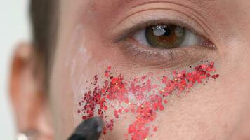 Extreme close-up view of make-up brush applying red sparkles to face of female model with brown eyes video